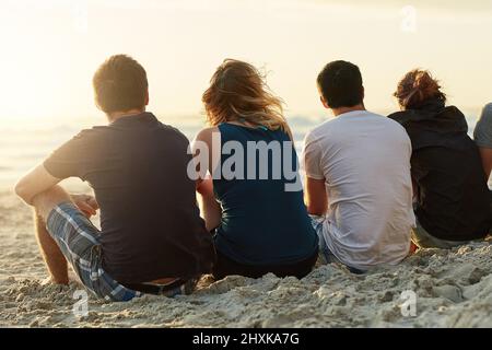 Trinken in der Aussicht zusammen. Rückansicht einer Gruppe von nicht identifizierbaren Freunden, die den Sonnenuntergang gemeinsam bewundern, während sie am Strand sitzen. Stockfoto