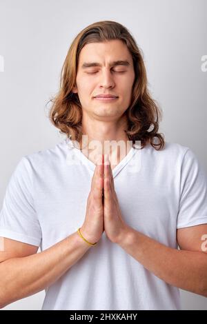 Ruhiger spiritueller hübscher Kerl, der mit geschlossenen Augen betet. Ernsthafter, friedlicher junger Mann, der sich den Händen anschließt, meditiert. Glaubenskonzept. kaukasischer Mann in Casu Stockfoto