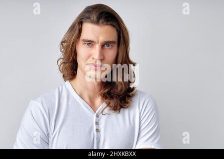 Der kaukasische Mann mit langen Haaren ist skeptisch und nervös und missbilligt den Gesichtsausdruck. Negative Person im lässigen weißen T-Shirt, posiert vor der Kamera. Mensch Stockfoto