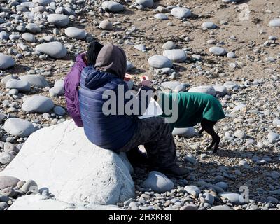 Ein Paar saß am Strand und aß Chips an einem kalten Frühlingstag, Westward Ho!, Devon, Großbritannien Stockfoto