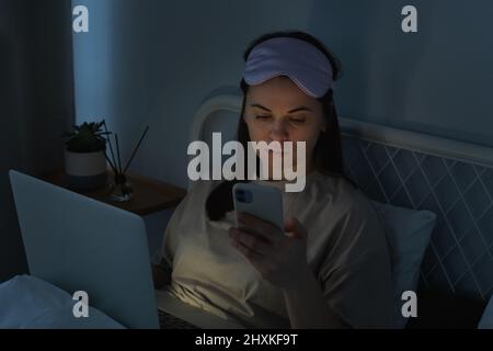 Frau in Seidenmaske mit Laptop-Computer und Surfen in sozialen Medien auf dem Smartphone im Bett vor dem Schlaf in der Nacht. Social-Media-Suchtkonzept Stockfoto