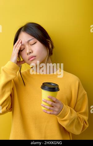 Frustrierte chinesische Frau leidet unter Kopfschmerzen, berührt Tempel, trinkt erfrischendes Getränk nach schlafloser Nacht, hält Einweg-Tasse Kaffee, dres Stockfoto