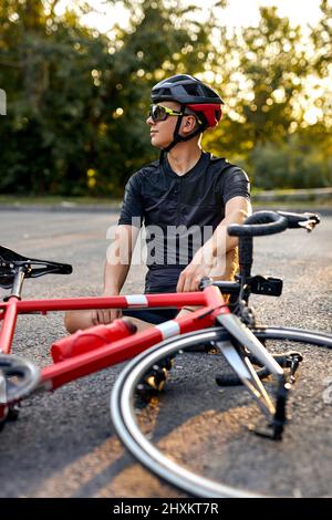 Sportlicher Typ Biker in Radbekleidung und Helm sitzen auf der Straße in der Nähe des Fahrrads, sich ausruhen. Mann, der sich nach dem Training auf dem Fahrrad in der Natur ausruhen und entspannen kann. Er Stockfoto