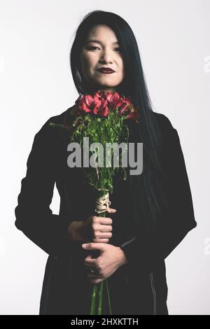 Beauty-, Lifestyle- und Modekonzept. Porträt einer asiatischen Frau mit langen schwarzen Haaren und Anzug in den Händen Blumenstrauß mit roten Blüten halten Stockfoto