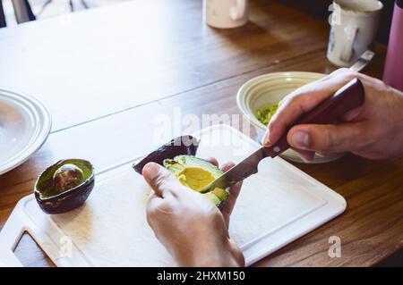 Rückansicht Nahaufnahme von männlichen Händen, die Avocado-Scheiben mit einem Messer auf einem weißen Schneidebrett auf einem Holztisch schneiden Stockfoto