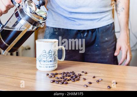 Mann, der zu Hause steht und Kaffee von einer italienischen Presse-Kaffeemaschine auf einem Holztisch neben gerösteten Kaffeebohnen in eine Porzellanbecher gießt Stockfoto