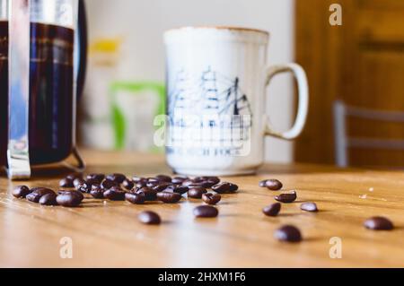 Kaffeebohnen auf einem Holztisch mit verschwommener Tasse und Kaffeekocher im Hintergrund in einer heimischen Küche Stockfoto