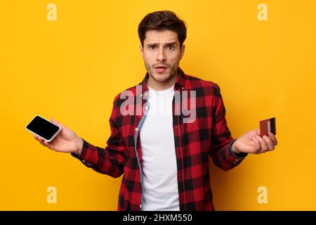 Verwirrt Mann hält Kreditkarte und Smartphone, zuckend Stockfoto