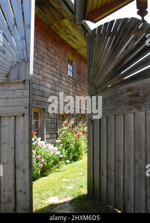 Hölzerne Scheunentüren führen zu einem alten Schindelhaus mit Blumenbeet im Vordergrund Stockfoto