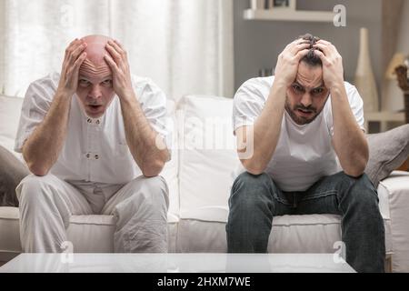 Älterer Vater und erwachsener Sohn, der ihren Kopf hält, sind erschrocken über schlechte Nachrichten oder negative Gedanken, die zu Hause auf der Couch sitzen Stockfoto