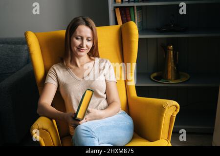 Porträt einer glücklichen blonden Frau, die in einem komfortablen gelben Stuhl sitzt und Papierbuch in einem dunklen Raum mit moderner Einrichtung hält. Stockfoto