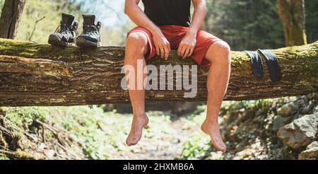 Mann, der auf einem umgestürzten Baumstamm über dem Bergwaldfluss sitzt, während er auf Socken wartet und Trekkingstiefel trocknet. Aktive Menschen auf Reisen, Stockfoto