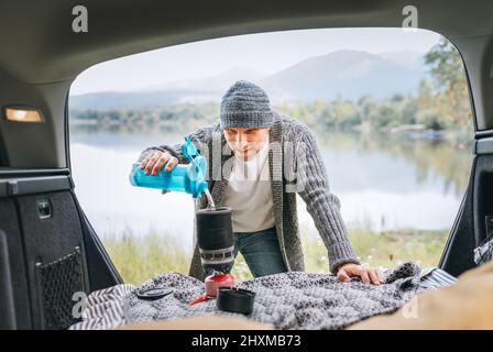 Ein lächelnder Mann in warmen Strickkleidern, der ein Frischwasser in den Gasherd in den gemütlichen Kofferraum mit einem wunderschönen Blick auf den Bergsee gießt. Warm Stockfoto