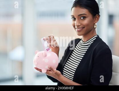 Selbst ein bisschen geht in der Wirtschaft einen langen Weg. Porträt einer attraktiven jungen Geschäftsfrau, die bei der Arbeit Geld in ihr Sparschwein steckt. Stockfoto