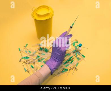 Weibliche Hand in einem schützenden Latexhandschuh, der die Spritze über einem gebrauchten Stapel infektiöser Nadeln hält Stockfoto