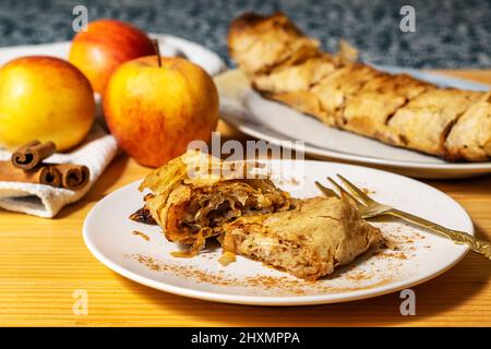 Eine Rolle hausgemachter frisch gebackener Apfelkuchen-Strudel mit einer appetitlichen Scheibe Apfelkuchen auf dem Teller und frischen Äpfeln und Zimtstangen auf einem hölzernen Stockfoto