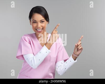 Stellen Sie sich Ihr Produkt hier vor. Beschnittenes Porträt einer attraktiven jungen Ärztin, die auf eine Kopiefläche im Studio gegen einen Grauen zeigt Stockfoto