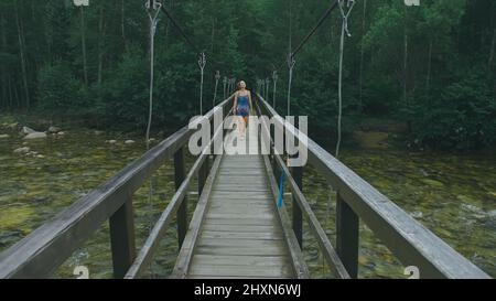 Frau, die alleine entlang der Hängebrücke in der malerischen Umgebung des grünen Waldes geht. Die Zeitlupe der dunkelhaarigen Frau geht auf der Hängebrücke im dichten grünen Wald. Keine anderen Menschen sind in the Shot. Stockfoto