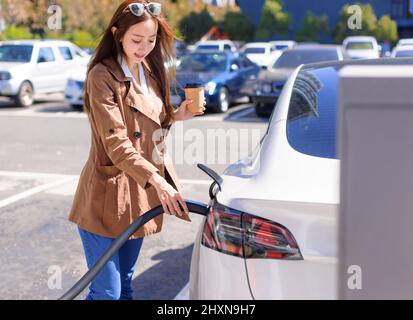 Lächelnde junge Frau, die auf dem Stadtparkplatz in der Nähe eines Elektroautos steht und die Autobatterie von einer kleinen Stadtstation lädt Stockfoto