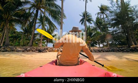 Junger Mann mit Sonnenbrille und Hut reiht sich mit einem rosa Plastikkanu entlang des Meeres gegen grüne, hügelige Inseln mit wildem Dschungel. Reisen in tropische Länder. Starker Kerl segelt auf Kajak im Ozean, Rückansicht. Stockfoto