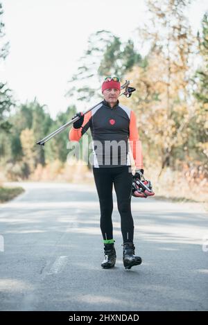 Training eines Athleten auf den Rollern. Biathlon-Fahrt auf den Rollskiern mit Skistöcken, im Helm. Herbsttraining. Rollensport. Erwachsener Mann, der auf Schlittschuhe reitet. Stockfoto