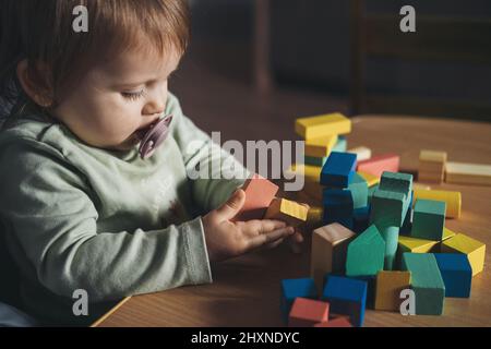 Babymädchen mit einem Schnuller im Mund spielt mit den bunten Figuren auf seinem Tisch. Kindererziehung. Gesunder Lebensstil. Entwicklung des Babys. Stockfoto