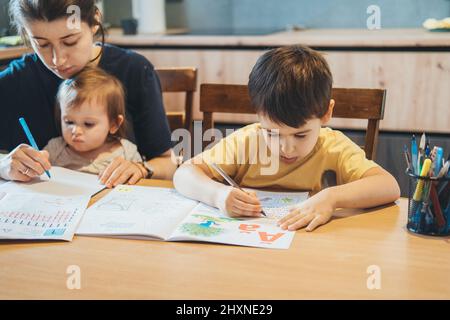 Junge, der am Tisch sitzt und das Schreiben praktiziert, Mutter hält das Schreiben eines kleinen Mädchens auf einem Laken konzentriert. Kindererziehung. Studieren, Hausaufgaben machen. Stockfoto