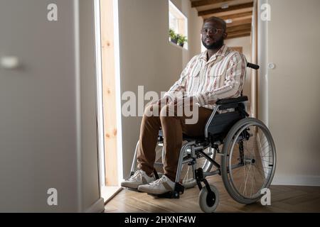 Ein junger, ernsthafter Mann mit Behinderung schaute auf die Kamera, während er im Rollstuhl vor der offenen Tür saß und nach draußen ging, um spazieren zu gehen Stockfoto