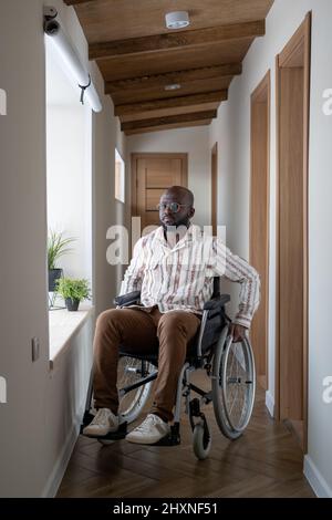 Afroamerikanischer Mann mit Behinderung sitzt auf dem Rollstuhl in einem langen Korridor einer großen modernen Wohnung und schaut auf die Kamera Stockfoto