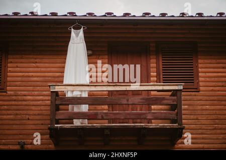 Weißes langes Hochzeitskleid, das auf einem Kleiderbügel auf dem Balkon eines Holzhauses hängt. Eleganter, moderner Stil. Elegantes Design. Sommerlicher Style. Stockfoto