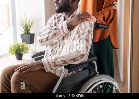Seitenansicht eines jungen afroamerikanischen Mannes mit Behinderung, der seine Hand auf die der kaukasischen weiblichen Betreuerin hält, die dahinter steht Stockfoto