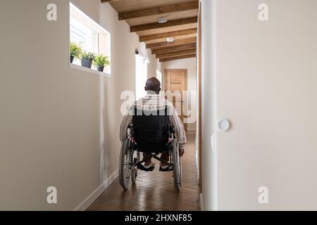 Rückansicht eines jungen afroamerikanischen Mannes mit Behinderung, der im Rollstuhl sitzt, während er sich auf dem Korridor zur Tür bewegt Stockfoto