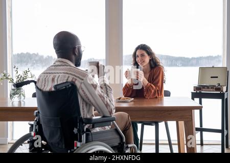 Fröhliche junge Frau mit Becher, die beim Küchentisch sitzt und beim Gespräch beim Frühstück auf einen schwarzen Mann mit Behinderung schaut Stockfoto