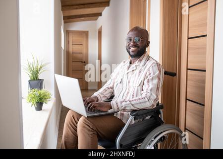 Junger erfolgreicher afroamerikanischer Geschäftsmann mit Laptop auf den Knien, der auf die Kamera schaut, während er auf dem Rollstuhl sitzt und sich mit ihm vernetzen kann Stockfoto