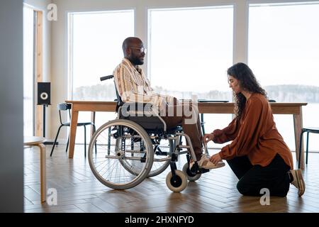 Junge weibliche Pflegekraft in der Freizeitkleidung, die Schnürsenkel eines afroamerikanischen Mannes mit Behinderung bindet, der im Wohnzimmer im Rollstuhl sitzt Stockfoto