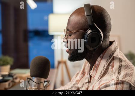 Seitenansicht eines jungen schwarzen Mannes in einem lässigen Hemd, Kopfhörern und Brillen, der seine Lieder singt und im Studio oder zu Hause aufnimmt Stockfoto