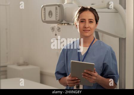 Glückliche junge, moderne Ärztin in blauer Uniform, die die Tablette hält, während sie gegen medizinische Geräte steht Stockfoto