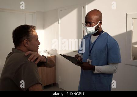 Reifer männlicher Patient, der sich über Schmerzen in der Schulter beklagt, und afroamerikanischer Arzt, der Notizen in einem medizinischen Dokument macht Stockfoto