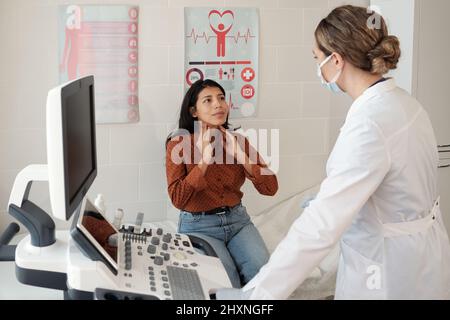 Junge hispanische Patientin berührt ihre Schilddrüse während der Selbstuntersuchung, während sie vor dem Arzt auf der Couch sitzt Stockfoto