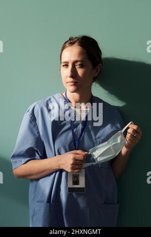 Junge Assistentin oder Ärztin in blauer Uniform mit Schutzmaske in den Händen, während sie an der Wand der Krankenhausabteilung steht Stockfoto