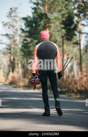 Training eines Athleten auf den Rollern. Biathlon-Fahrt auf den Rollskiern mit Skistöcken, im Helm. Herbsttraining. Rollensport. Erwachsener Mann, der auf Schlittschuhe reitet. Stockfoto
