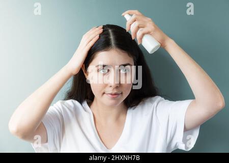 Porträt einer kaukasischen Frau mit dunklem Haar, die das Sprühen eines kosmetischen Produkts auf den Haarscheitel anwendet. Grüner Hintergrund. Das Konzept der Haarpflege und da Stockfoto