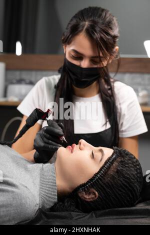 Frau tätowiert permanent Lippen Make-up im Schönheitssalon, Lippenvergrößerung durch Auftragen von Pigment, Kosmetikerin macht Make-up Verfahren mit Tattoo-Maschine Stockfoto