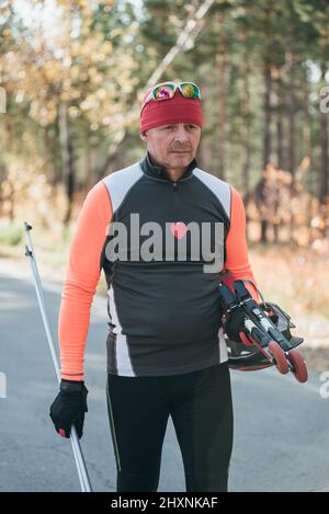 Training eines Athleten auf den Rollern. Biathlon-Fahrt auf den Rollskiern mit Skistöcken, im Helm. Herbsttraining. Rollensport. Erwachsener Mann, der auf Schlittschuhe reitet. Stockfoto