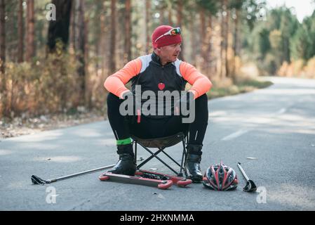 Training eines Athleten auf den Rollern. Biathlon-Fahrt auf den Rollskiern mit Skistöcken, im Helm. Herbsttraining. Rollensport. Erwachsener Mann, der auf Schlittschuhe reitet. Der Athlet bereitet sich auf den Start vor. Stockfoto