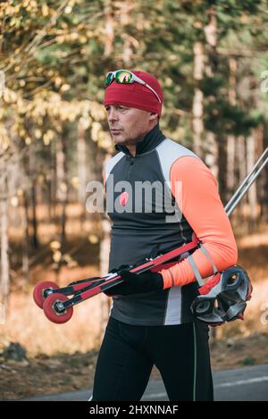 Training eines Athleten auf den Rollern. Biathlon-Fahrt auf den Rollskiern mit Skistöcken, im Helm. Herbsttraining. Rollensport. Erwachsener Mann, der auf Schlittschuhe reitet. Stockfoto