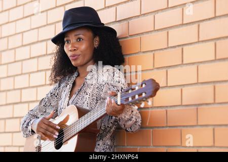 Beschnittene Ansicht einer Musikerin auf der Straße, die sich an einer Backsteinmauer lehnt und Gitarre spielt Stockfoto