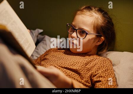 Portrait eines jungen Mädchens mit Down-Syndrom, das nachts im Bett ein Buch liest, das von einem gemütlichen Nachtlicht beleuchtet wird Stockfoto