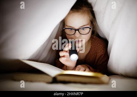 Vorderansicht Porträt eines jungen Mädchens mit Down-Syndrom, das Buch unter Deckeln mit Taschenlampe liest, Kopierraum Stockfoto