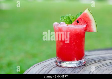 Ein Glas Wassermelone-Smoothie wird für das Sommergetränk-Konzept auf einen Tisch mit verschwommenem grünem Gartenhintergrund gestellt. Stockfoto